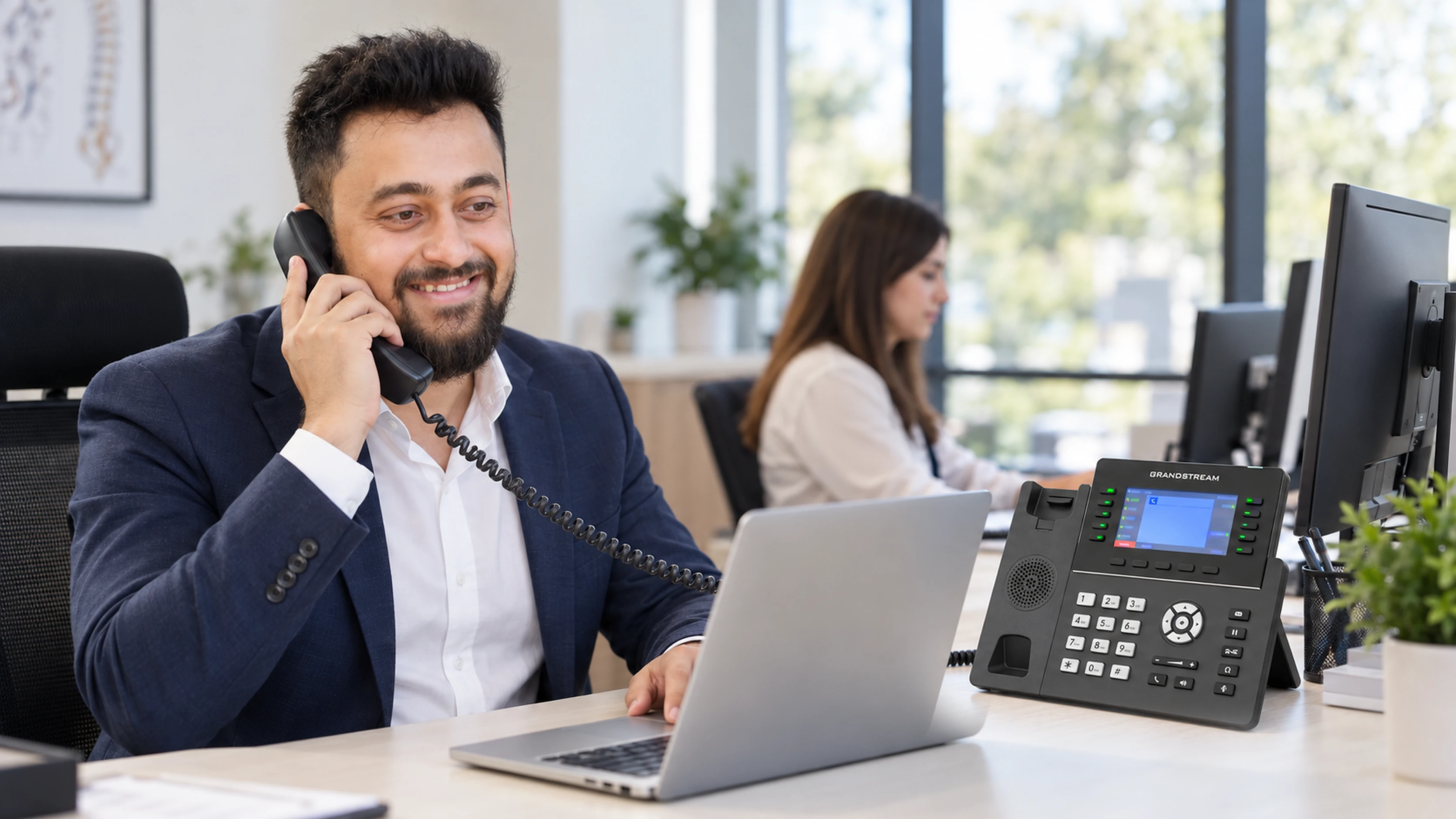 A man at an office desk taking a call on a handset, a laptop open in front of him and a GRP2615 IP desk phone beside it